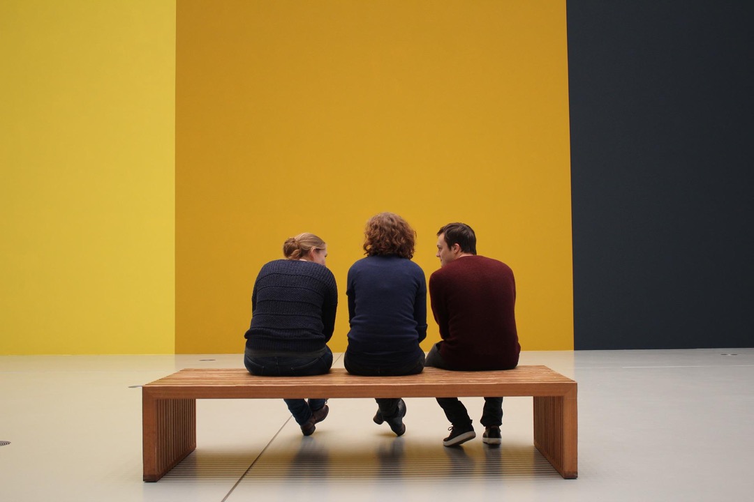 Foto van drie mensen op een bankje in een ruimte die er als museum uitziet.